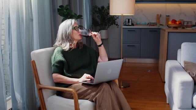 Mature woman drinks wine while using laptop in armchair at home