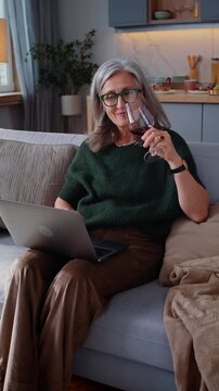 Aged woman enjoys wine using laptop on free time on sofa at home