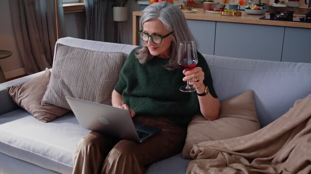 Mature grey-haired woman reads laptop content holding wineglass