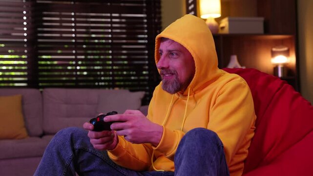 Emotional man with controller playing video game on pouf at home