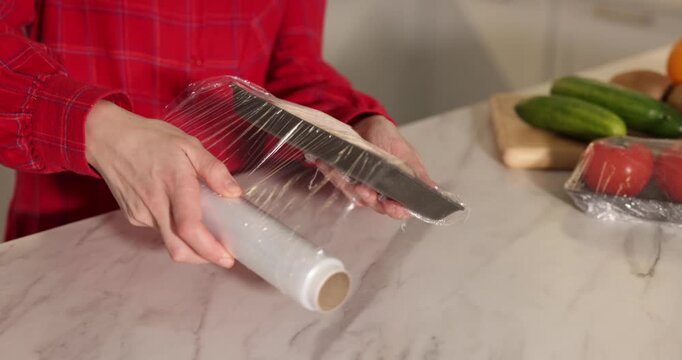 Woman wrapping container with raw chicken wings in cling film at light marble table, closeup