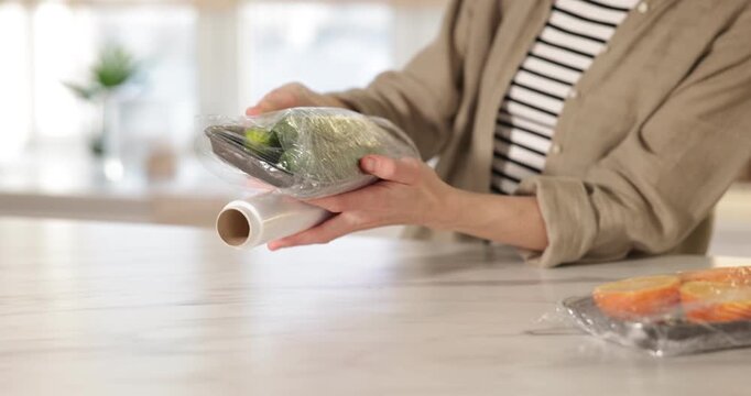 Woman wrapping container with fresh cucumbers in cling film at light marble table, closeup