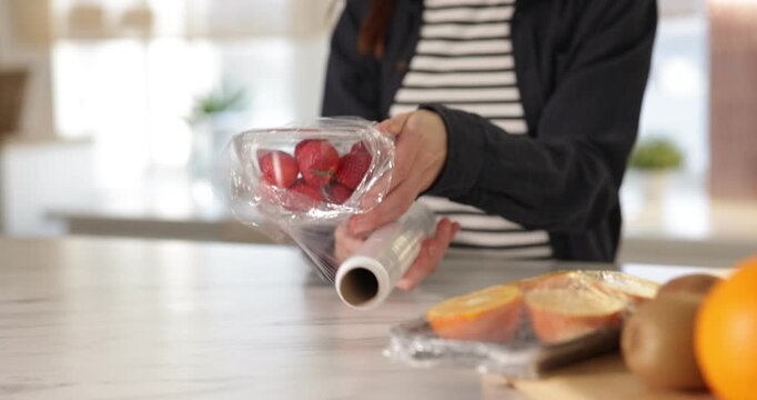 Woman wrapping bowl with fresh strawberries in cling film at light marble table, closeup