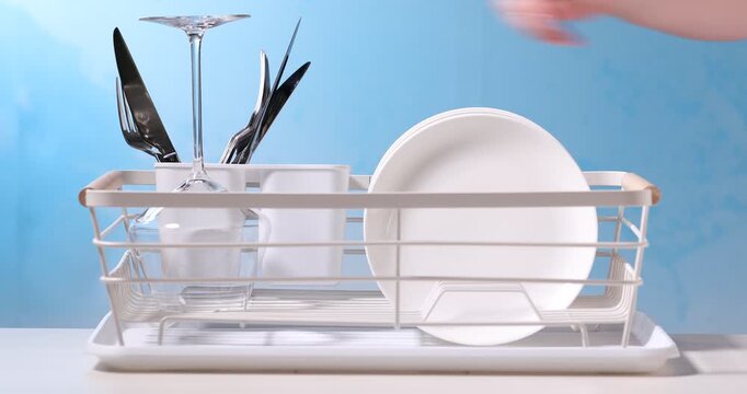 Woman putting clean cutlery and glasses into dish rack on white table against light blue background, closeup
