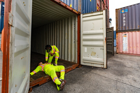 Logistics worker providing first aid to an unconscious colleague in a cargo container. Emergency response and occupational safety training for industrial accidents at a port terminal.