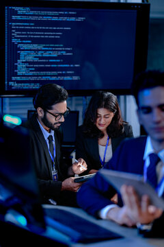 An AI research team brainstorms late night. Innovators discuss machine learning model architecture using a tablet while reviewing training data code displayed on a large screen.