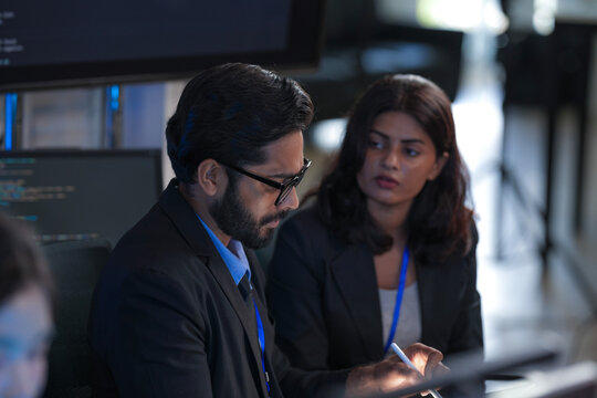 Focused diverse professionals collaborate late night in a modern tech office. Colleagues discuss project details using a tablet with stylus while code runs on a large screen.