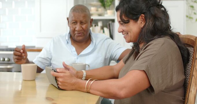 Senior couple at kitchen table, woman tapping tablet while man with mug looking up and gesturing