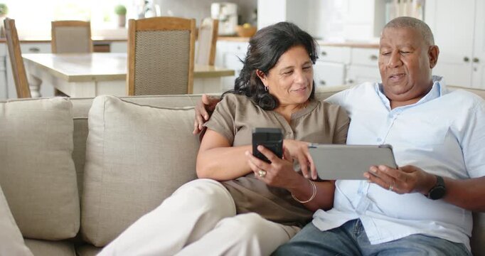 African American senior couple on couch at home, man tilting tablet while woman comparing phone
