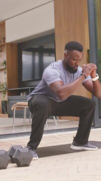 Vertical video: Arriving mat man in grey tee lowering into squat on patio with hex dumbbells left