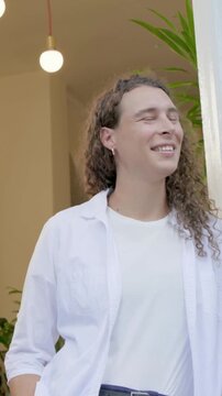 Vertical video: Camera moving, adult leaning in doorway, white shirt and plants, smiling, greeting