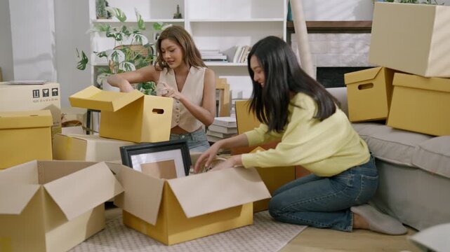 Moving packing cardboard box apartment friend woman relocation new home home interior unpacking Two young adult women packing and unpacking household items into cardboard boxes apartment living room