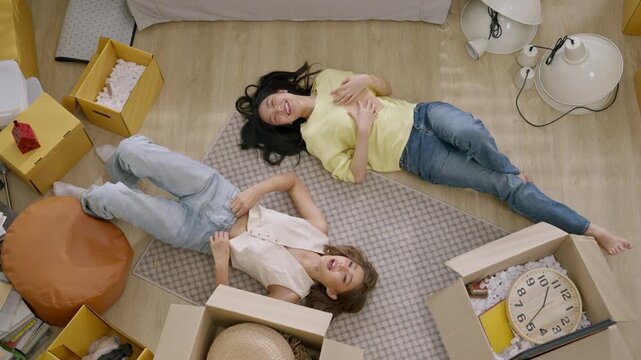 Young woman and female friend relaxing on floor with moving box in new home apartment, packing and unpacking from cardboard box in home interior, overhead view, friendship during moving day