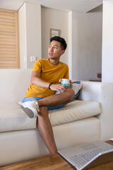 Adult Asian man sitting on white sofa holding teal-and-white mug, silver laptop on coffee table © wavebreak3