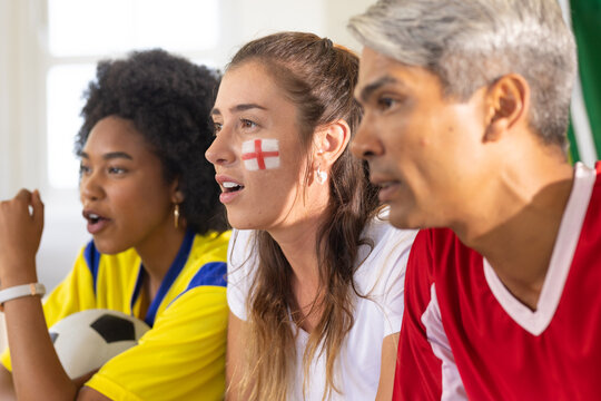 Diverse fans watching soccer match on couch, in jerseys, holding ball, showing red cross paint