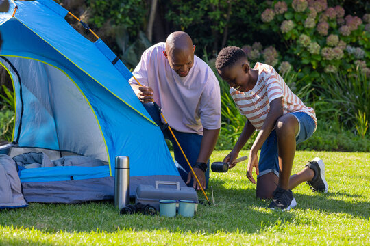 African American dad and youth son using mallet, adjusting yellow pole, prepping blue tent in yard