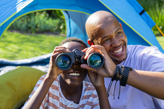 Vertical video: Father and son looking for birds with binoculars in sunny backyard at blue tent