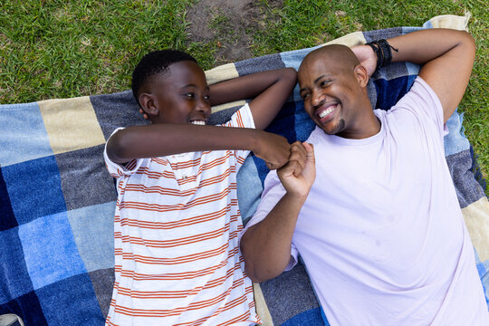 African American father and son lying on picnic blanket on grass, fist-bumping, wristband visible
