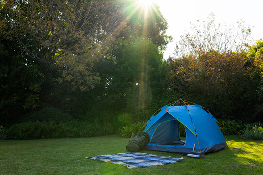 Blue dome tent on mown lawn sun beaming, revealing blanket, green pack leaning, thermos, copy space