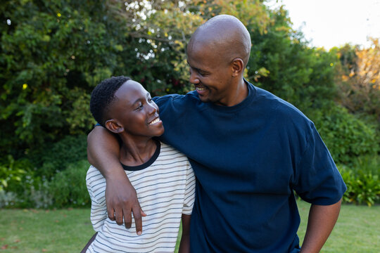 African father in blue shirt draping arm around youth in striped tee on grassy lawn, smiling
