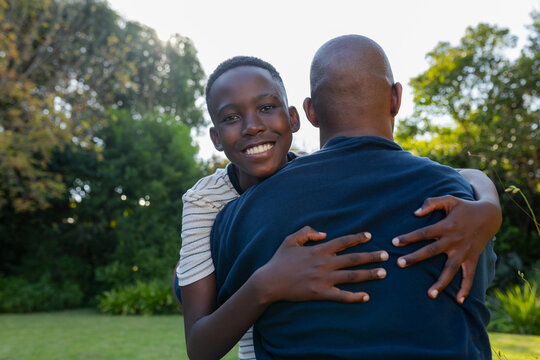 African dad and youth, youth hugging dad from behind after spotting camera, posing on grassy lawn