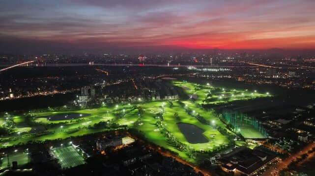 Detailed aerial sunset view of a golf course in Long Bien district, Hanoi, showing illuminated fairways, practice range, water hazards, and expansive surrounding cityscape.