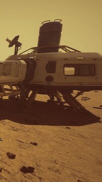 A habitat module is set on the surface of Mars with rocks scattered around. The environment shows a dusty landscape with a hazy sky. The setting sun casts an orange glow on the scene.