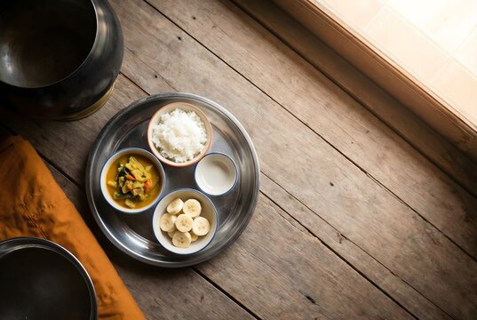 Top-down flat lay of a monk's simple morning meal arranged on a traditional Thai offering tray &mdash; rice, curry, fruit &mdash; with edge of saffron robe and alms bowl visible, warm natural window light.