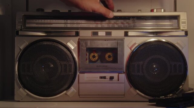 Detailed close-up: hand inserts cassette tape into classic boombox on table under soft warm light, then presses the play button. Tape starts playing old music, nostalgic vintage moment 4K.