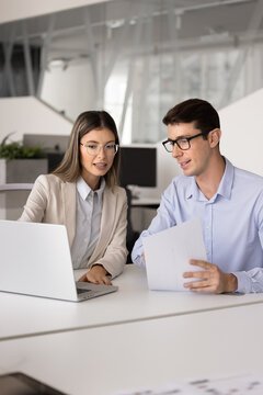 Two young professionals collaborating at desk in modern office, businesswoman pointing at laptop screen while businessman holds paper, analyzing data and discuss project strategy, engaged in teamwork