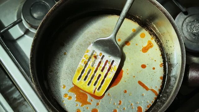 Overhead close-up of a dirty frying pan with metal spatula and orange oil residue on a stovetop, showing post-cooking mess, greasy surface, and used cookware in an indoor kitchen setting.