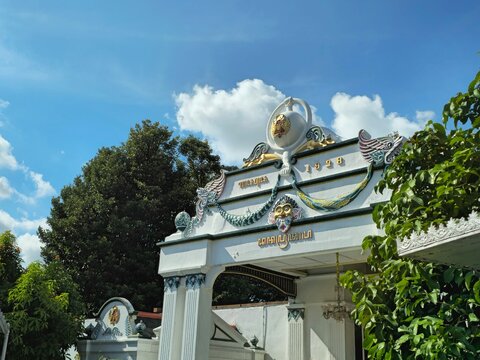 Main courtyard entrance at Keraton Yogyakarta with white ornate gate, wide paved yard, surrounding traditional buildings, potted plants, trees, and clear blue sky above.