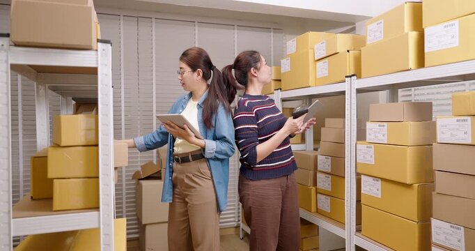 Asian women online business partners review inventory shelf together, checking parcel box quantity, confirming order readiness, supporting organized e-commerce warehouse workflow inside home storage