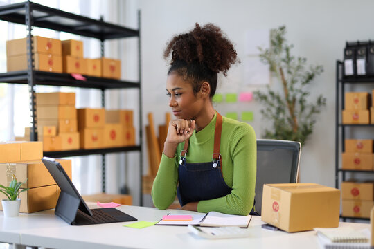 Young African American woman business owner using digital tablet checking ecommerce orders of her online store in warehouse, selling online concept.