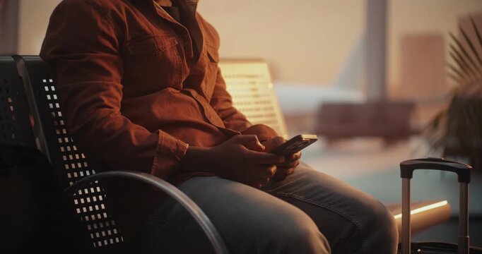 Man in Stylish Overshirt Sits on Terminal Bench, Fully Engaged With Smartphone and Types Message. Everyday Moment of Traveler Staying in Touch With Loved Ones, Sharing Updates During Flight Layover.