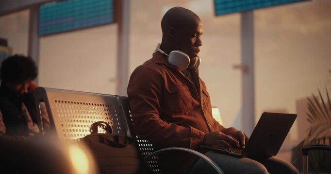 African American Man Sits on Terminal Bench, Deeply Focused on Laptop Screen While Waiting For Flight. Concept of Freelance Lifestyle, Corporate Travel, Mobile Office Solutions, Global Connectivity.