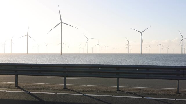 Wide roadside view of passing cars with an offshore wind farm in soft morning light.