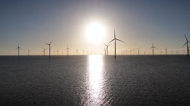 Aerial forward view approaching the wind farm with sun reflections shimmering across the water.