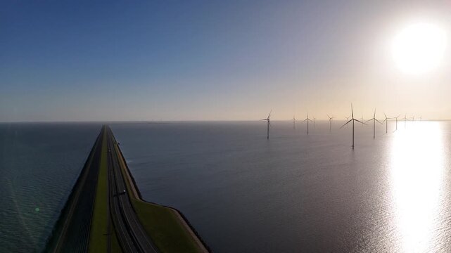 Backlit aerial shot toward the distant curve of the dam with wind turbines and bright reflections across the water.