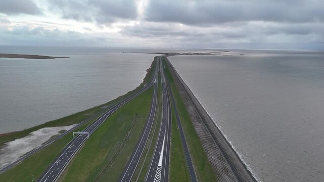 Drone tracking shot above the roadway as the wide approach narrows into the long Afsluitdijk under overcast evening light.