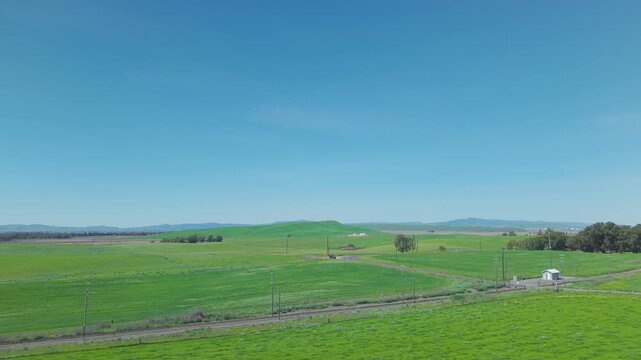 Aerial footage moving sideways over a vast green agricultural landscape with rolling hills, a railway line, and a clear blue sky in rural Birds Landing, Solano County, California, USA
