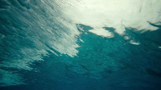 Underwater ocean surface with bright sunlight filtering through water, pan follows motion of wave crashing and breaking naturally
