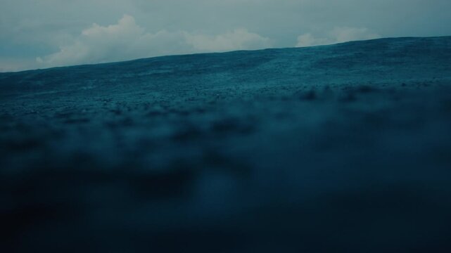 Low angle across stormy raindrops on ocean surface toward distant horizon as wave rises and bump crosses fading to dark, slow motion textured water backdrop