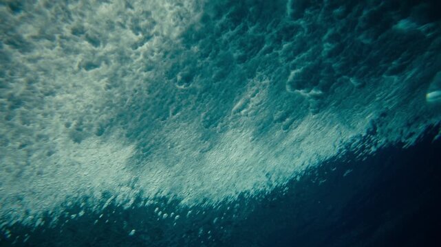 Underwater perspective looking up at smooth blue ocean wave surface as wave lip crashes spreading mist and vortex behind curling barrel, slow motion