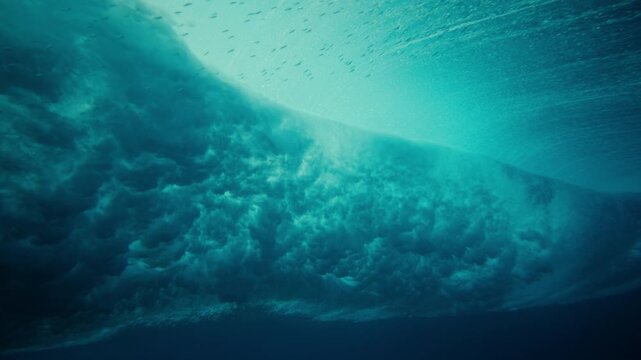 Underwater of powerful breaking ocean wave with turbulent bubbles and blue water, vortex barrel and fish swimming at surface sparkling in slow motion