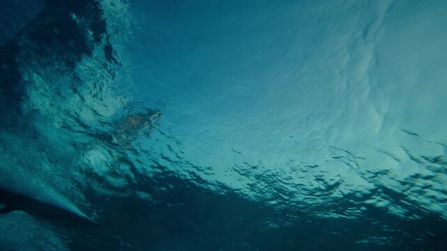 Underwater ocean beneath wave with dark blue water and surface ripples, rearview follows shimmering surfer silhouette riding wave, behind pov