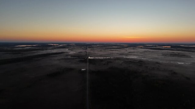 High aerial drone shot panning right over boreal forest in northern Manitoba before sunrise. Train tracks ahead with War Lake Ilford First Nation in distance under dark sky and misty forest.