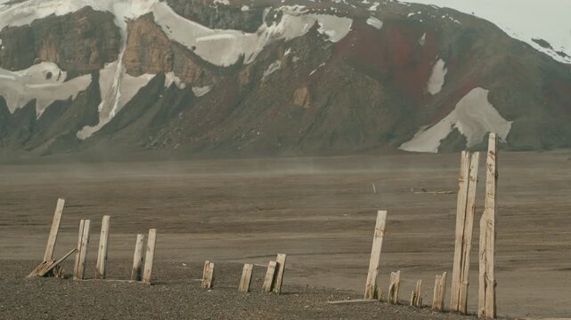 Weathered wooden planks of abandoned whaling station ruins stand on dark volcanic ash plain at Whalers Bay, Deception Island, with snow-capped volcanic mountains behind