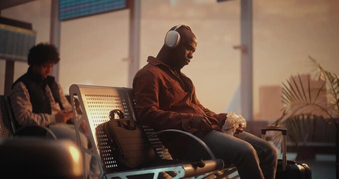 African American Man Sits on Bench in Airport Departure Lounge, Bathed in Warm Sunset Glow From Panoramic Windows. Male Wearing Headphones and Holds Hamburger, Enjoying Quiet Meal During Transit.