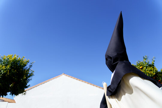 Nazareno penitent in a traditional black Pointed hood and tunic holds a candle while participating in a solemn holy week procession, representing religion and devotion under a clear blue sky